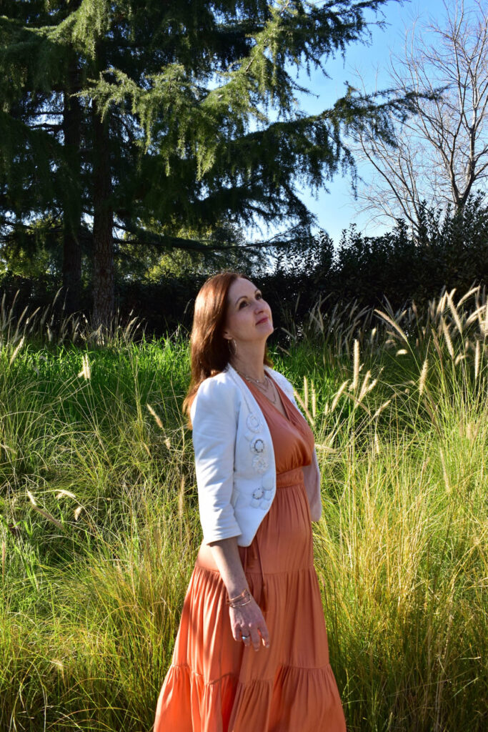 Simona standing in a field wearing a light coral sundress and white coat